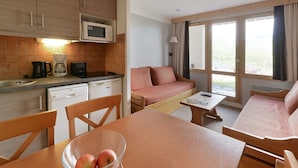 Habitación de hotel compacta con kitchenette, sofá, mesa de comedor y un amplio ventanal con vistas al exterior.