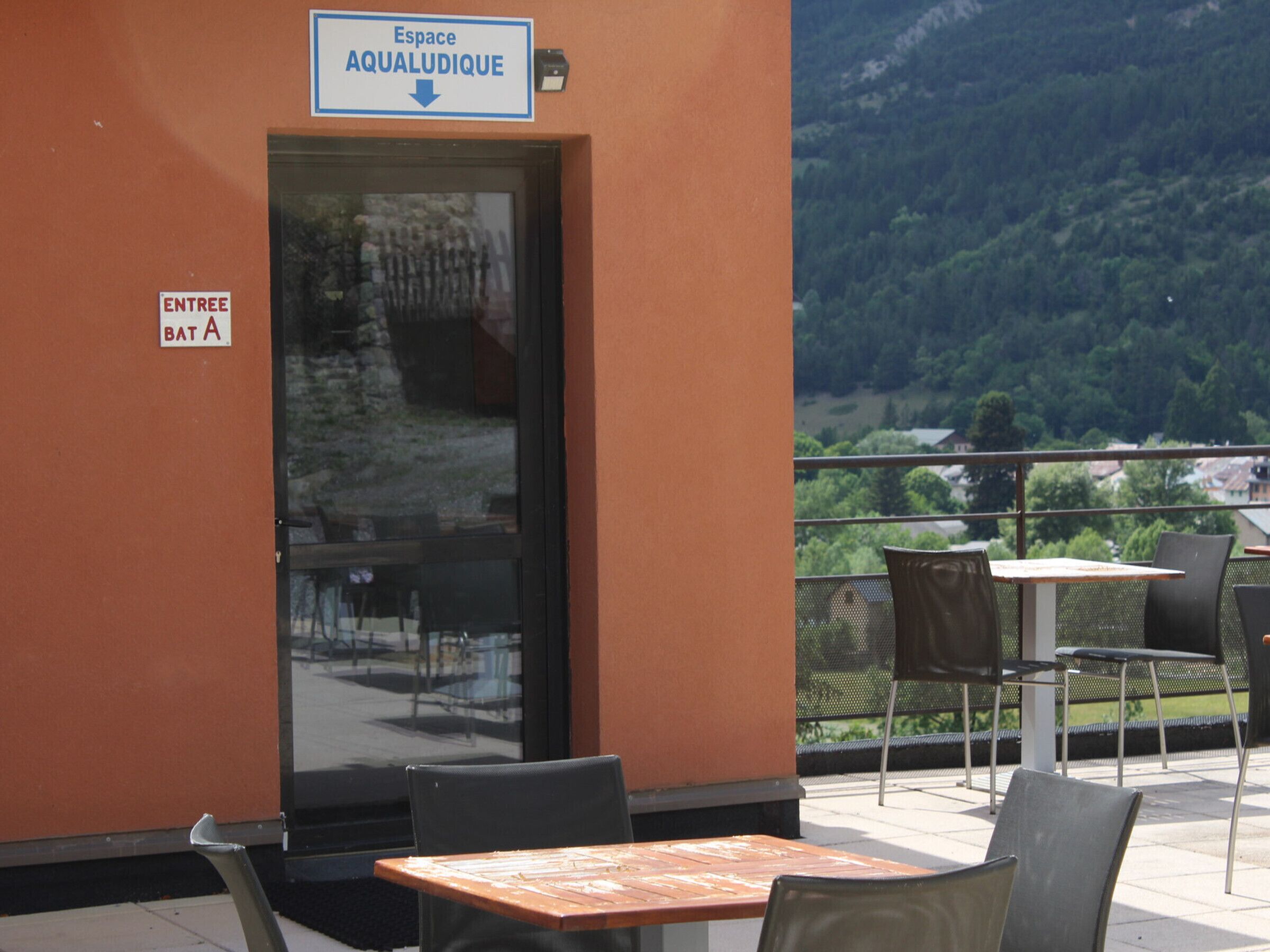 An outdoor seating area with tables and chairs, a door labeled 'ENTREE BAT A,' and a sign indicating 'Espace AQUALUDIQUE.'