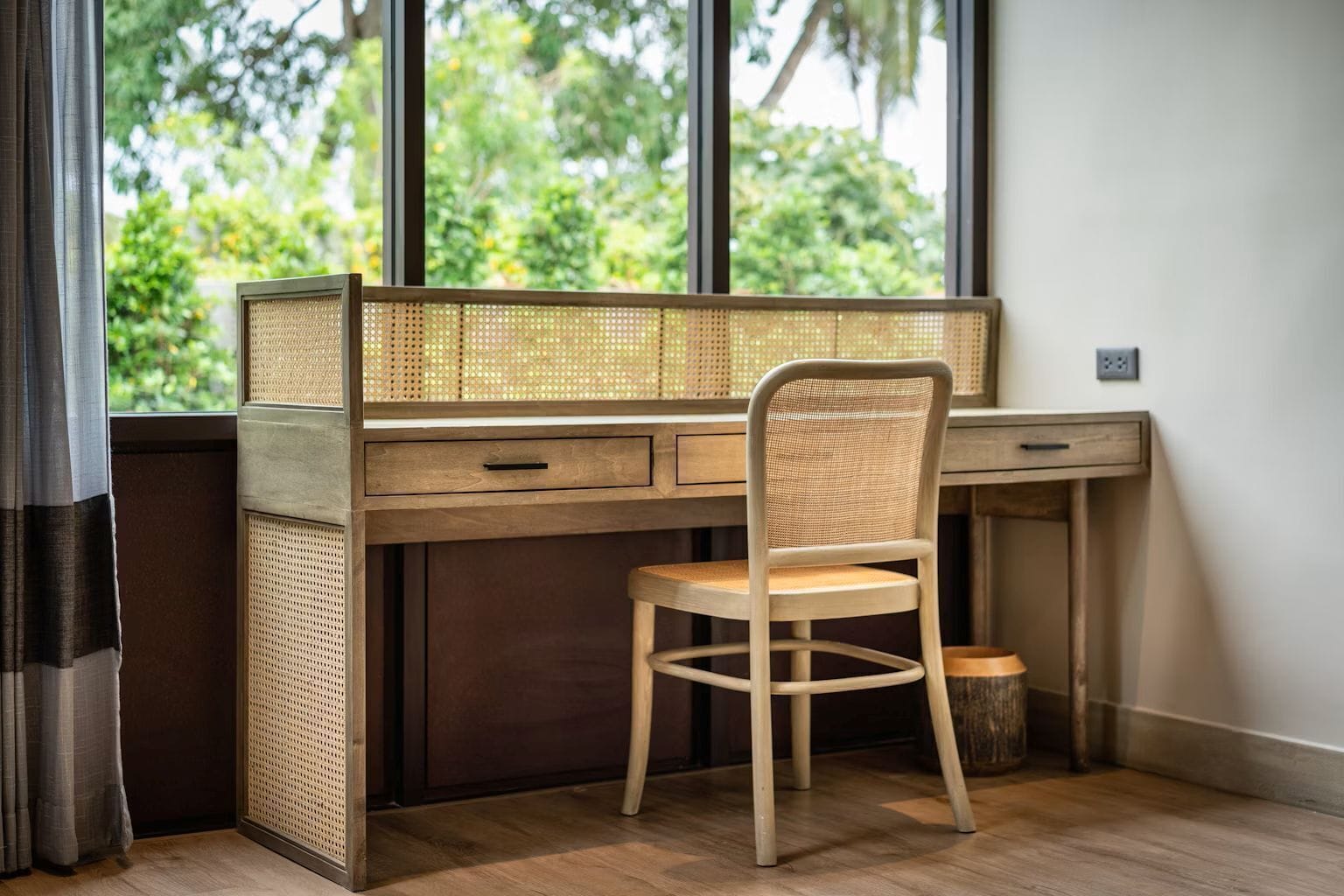 A workspace with a wooden desk, a wicker chair, and a view of greenery through the window.