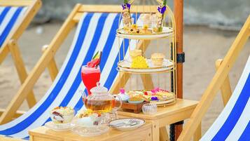 A beachside lounge chair with a tiered dessert stand, a glass of red beverage, and various snacks and pastries.