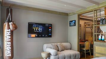 A modern hotel room with a grey sofa, a punching bag, a flat-screen TV, and a wooden shelf with bottles.