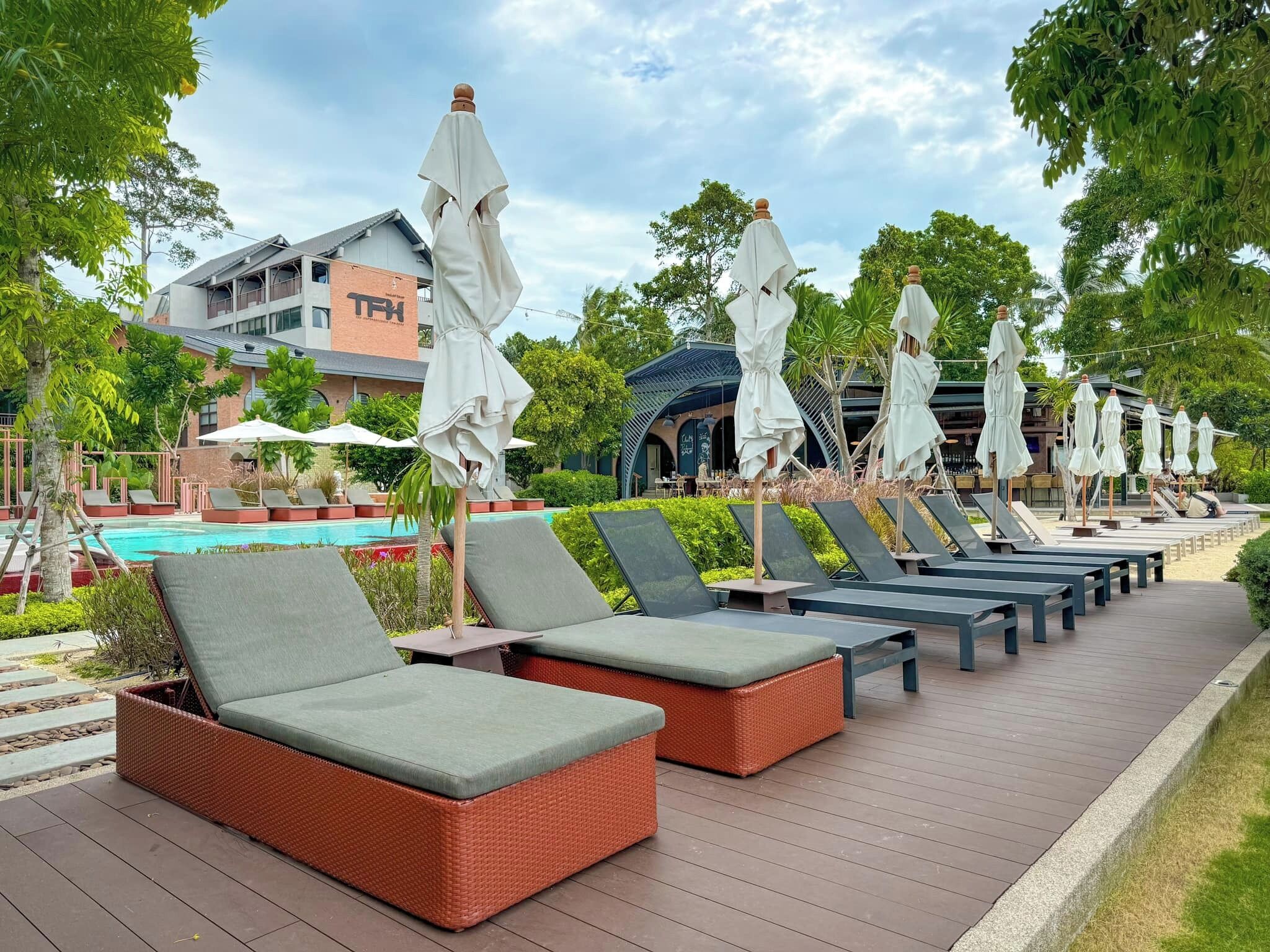 A row of sun loungers with umbrellas, a pool, and a building with a sign in the background.