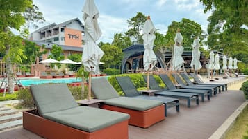 A row of sun loungers with umbrellas, a pool, and a building with a sign in the background.