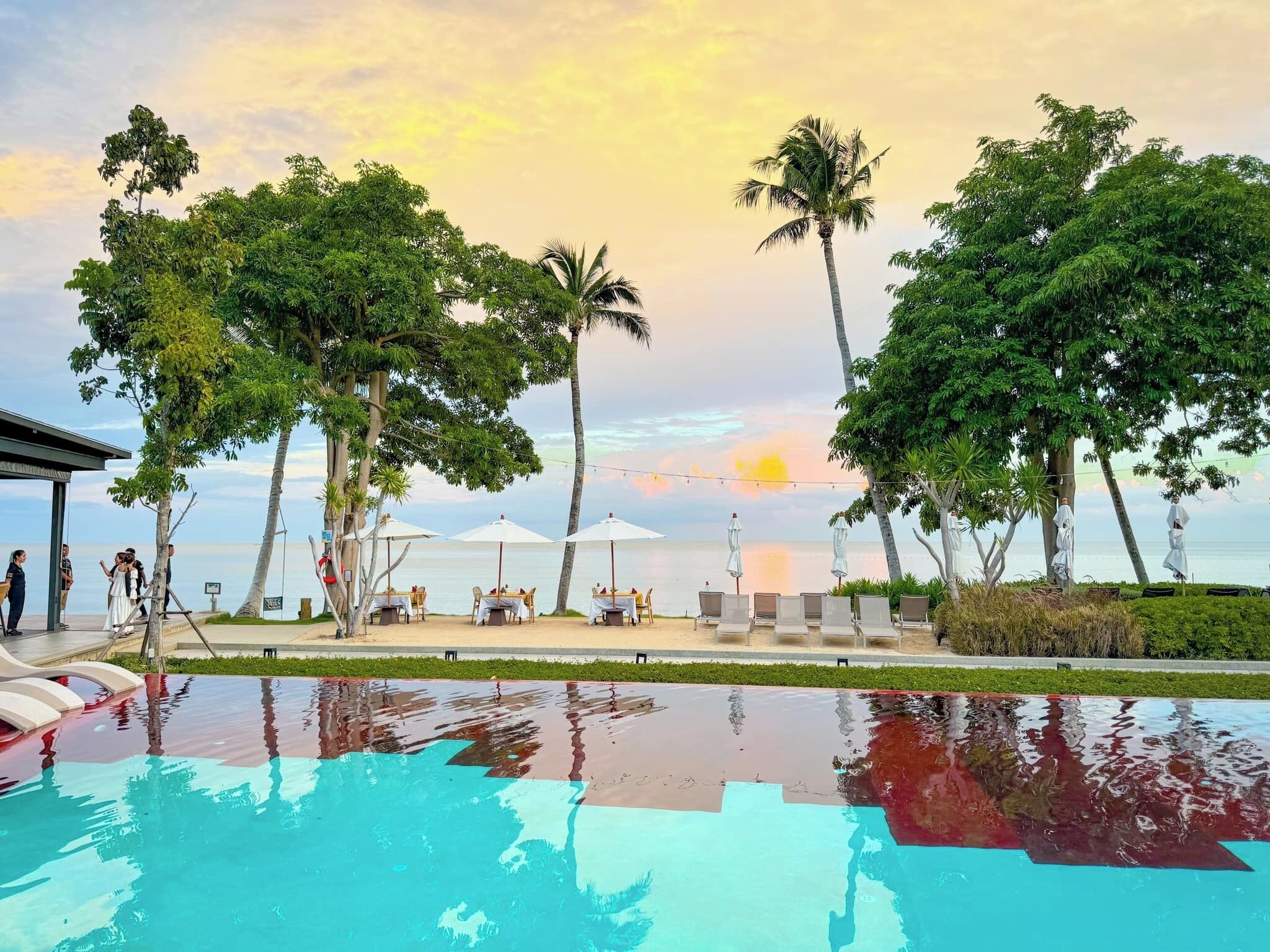 Ein Poolbereich mit Sonnenuntergangsblick, umgeben von Palmen und Strandmöbeln.
