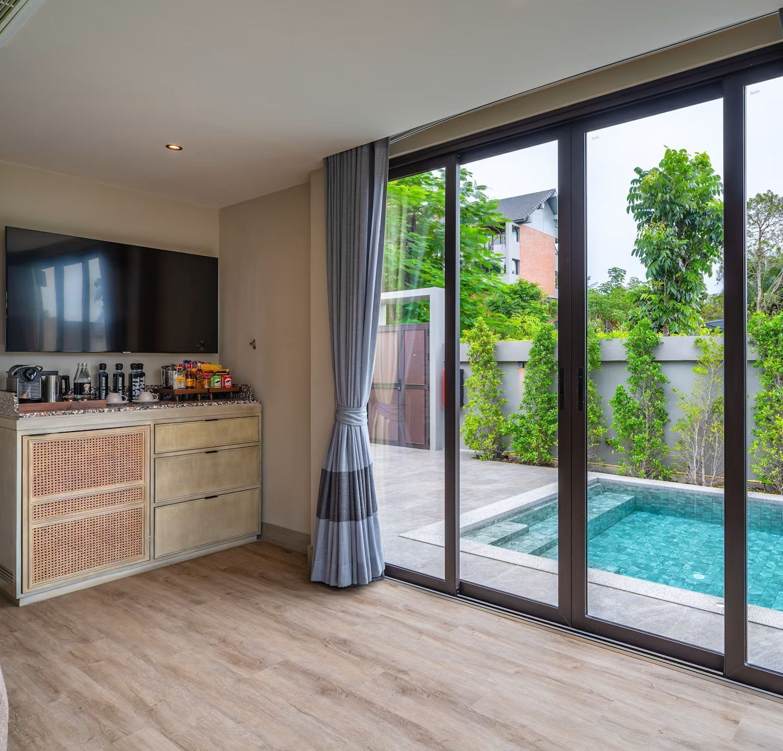 Una sala moderna con televisor de pantalla plana, una encimera de cocina con varios objetos, una gran puerta corrediza de vidrio que da a una piscina y vistas a la vegetación exterior.
