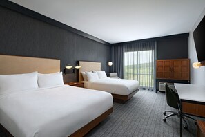 Quarto de hotel com duas camas, uma escrivaninha e uma cadeira. Há duas luminárias na parede e uma janela grande com cortinas.