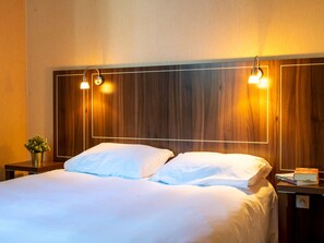 Une chambre d’hôtel avec une tête de lit en bois, deux lampes de chevet, un lit avec du linge de lit blanc, une petite table de chevet avec une plante et une table de nuit avec des livres.