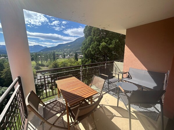 A balcony with a wooden table and chairs, a patterned tablecloth, and a view of a mountainous landscape.