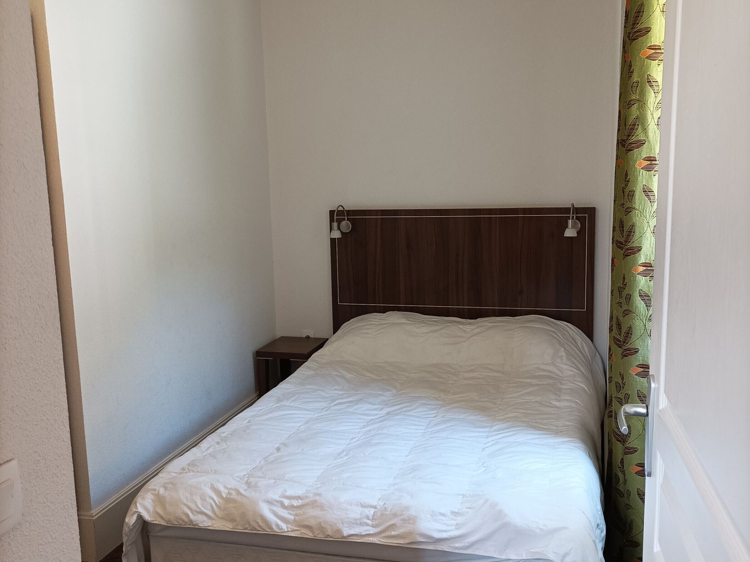 A small, narrow room with a single bed, a nightstand, and a wooden headboard.