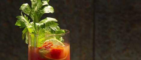 Un vaso de Bloody Mary con apio y una cuña de lima, sobre una superficie reflectante y un fondo oscuro y con textura.
