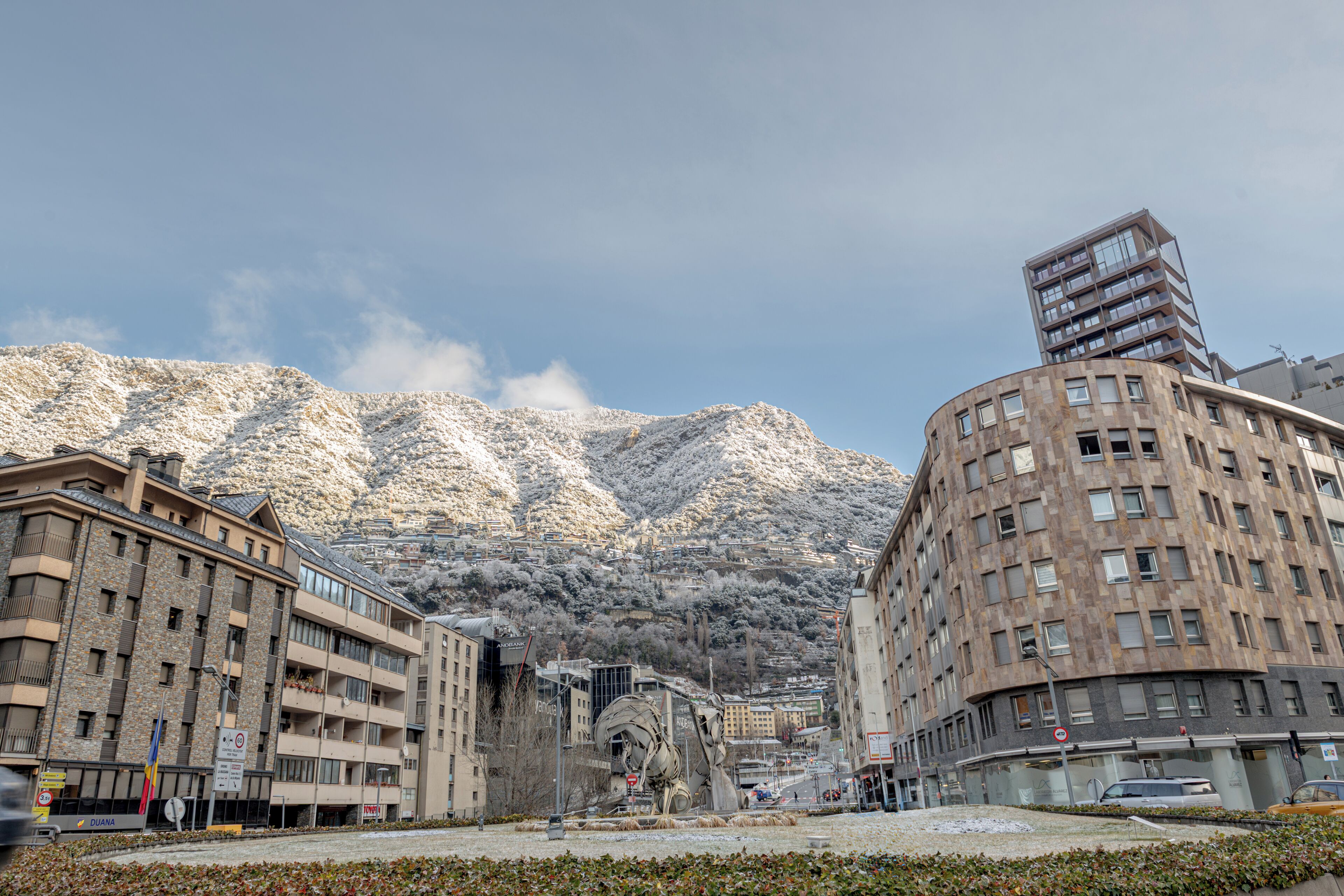 Un paisaje urbano con montañas nevadas al fondo, un edificio moderno de diseño único, y un edificio tradicional de varios pisos.