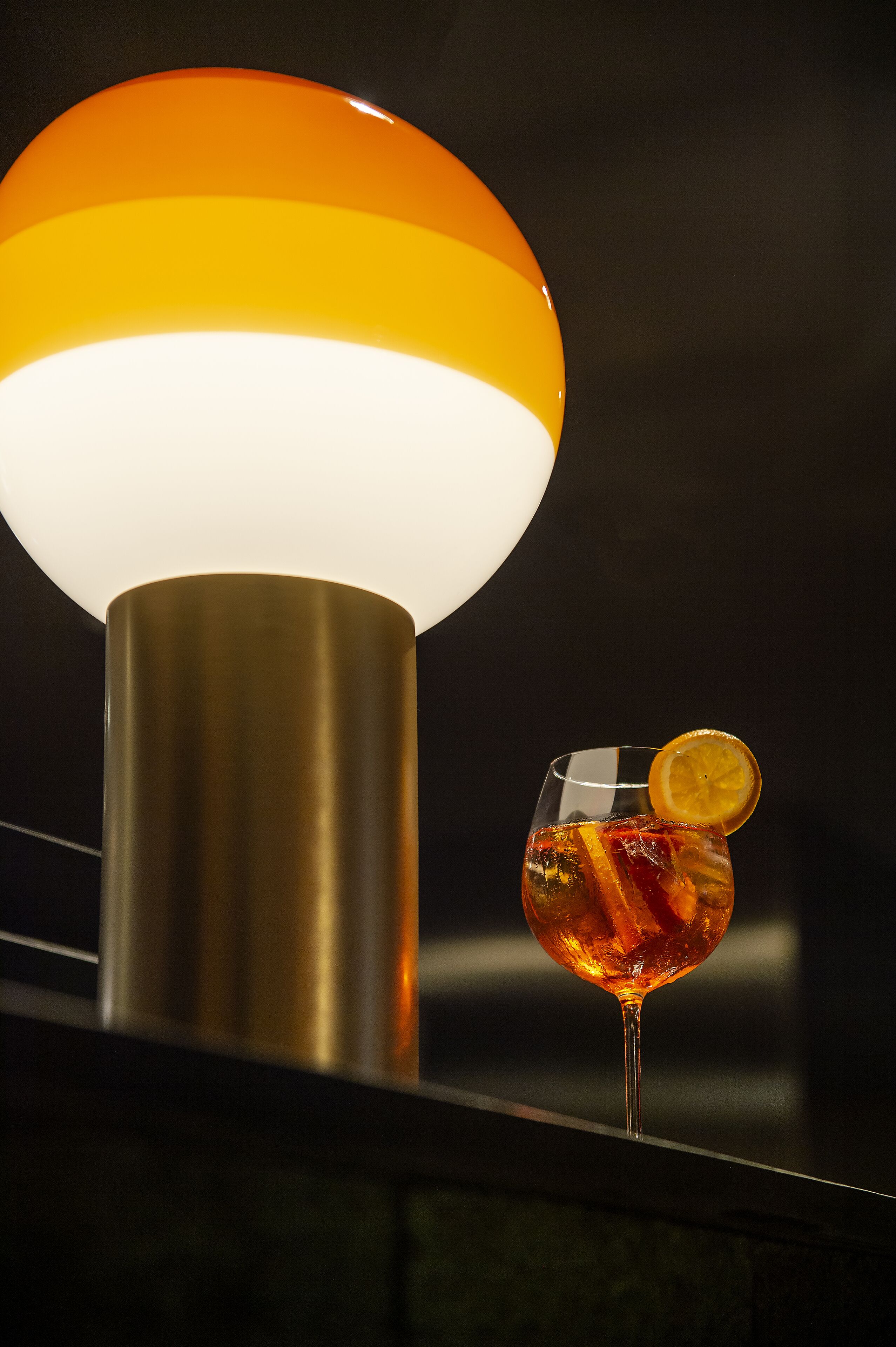 A cocktail glass with a lemon slice garnish on a bar counter, next to a modern lamp with a metallic base and a glowing orange light.