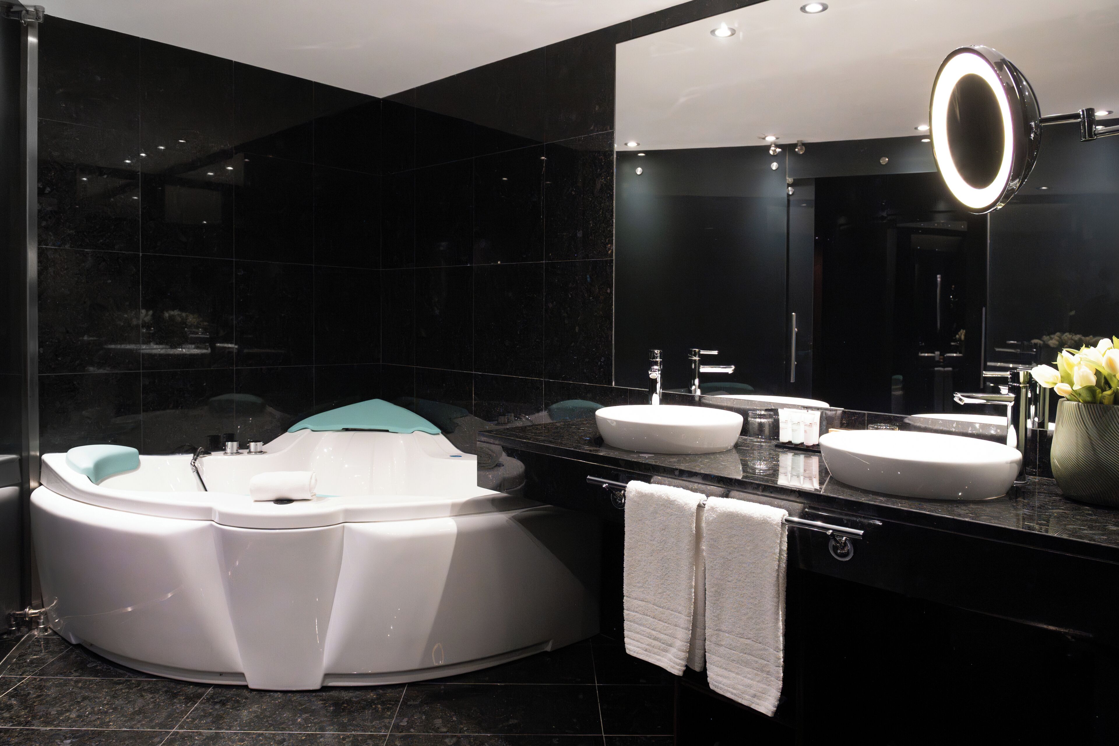 A modern bathroom with a jacuzzi, two sinks, a large mirror with a light, and a vase with flowers.