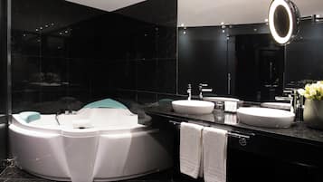 A modern bathroom with a jacuzzi, two sinks, a large mirror with a light, and a vase with flowers.