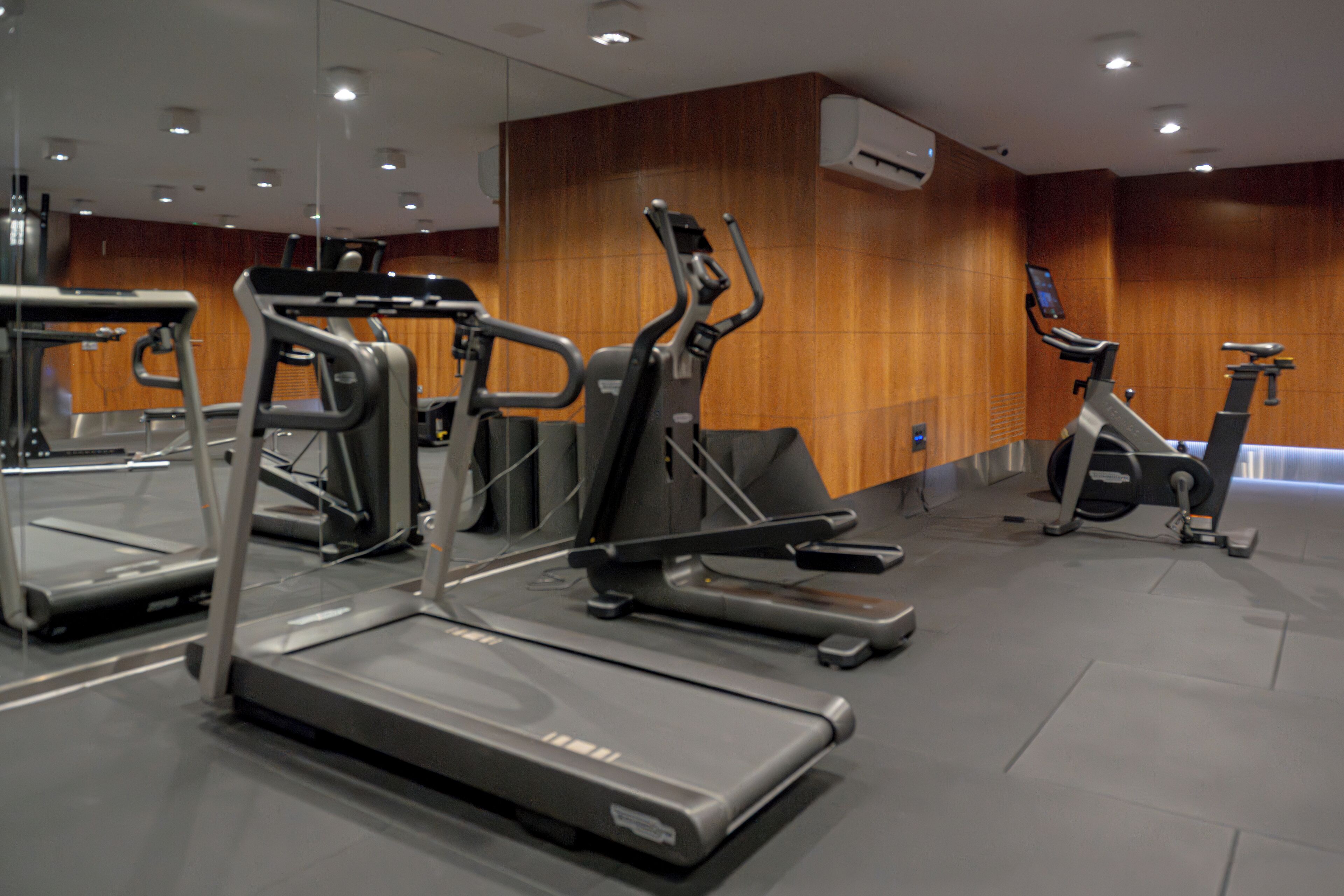 Ein Fitnessraum mit mehreren Laufbändern und Crosstrainern, Holzwandverkleidung und einer Spiegelwand.