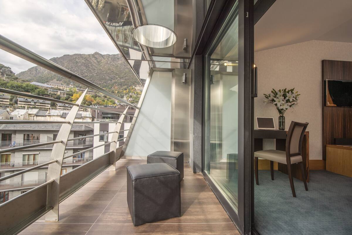 Un balcon avec vue sur les bâtiments et les montagnes, une table avec une chaise et un vase rempli de fleurs.