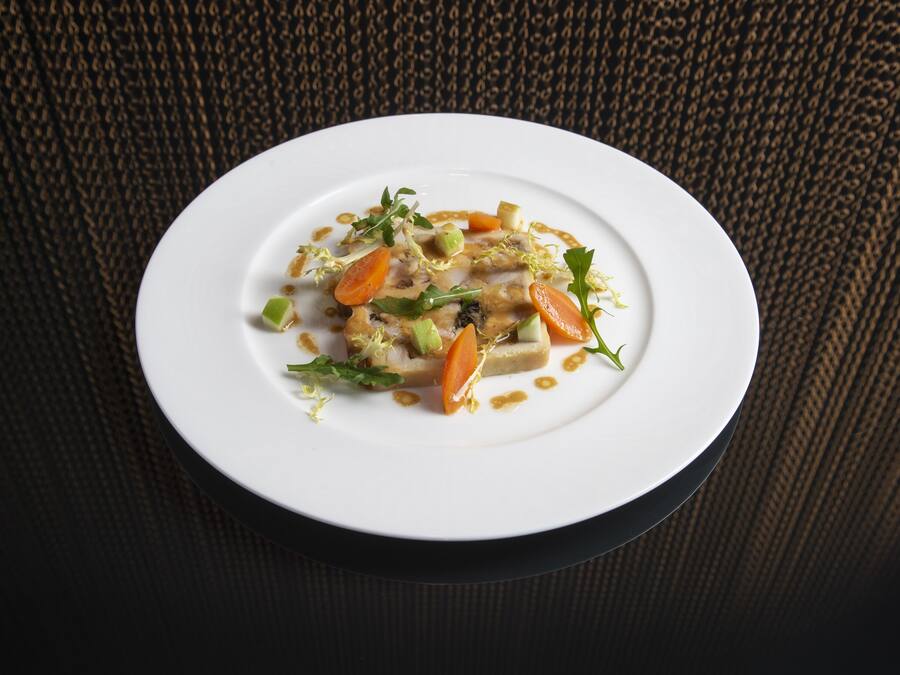 A white plate with a dish of food, including vegetables and a sauce, on a textured dark background.