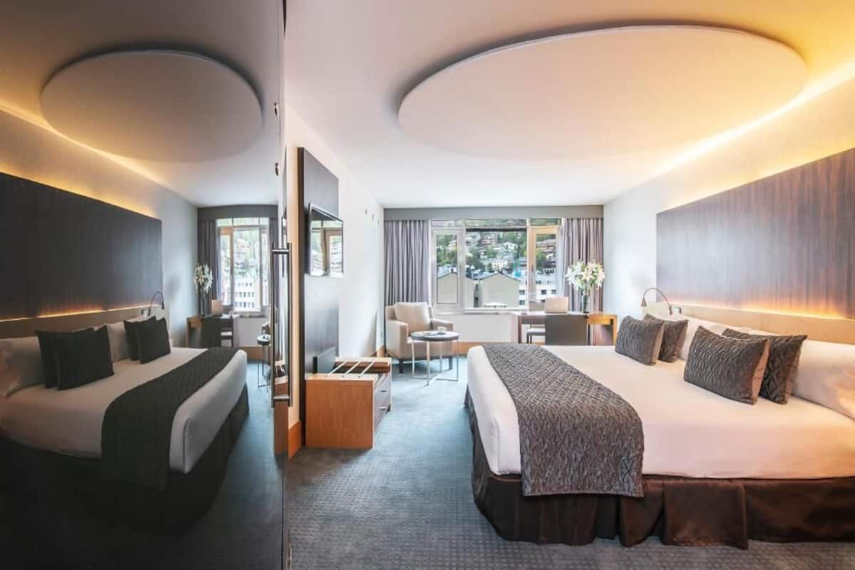 A hotel room with two beds, a desk, a chair, and a large window with a city view.