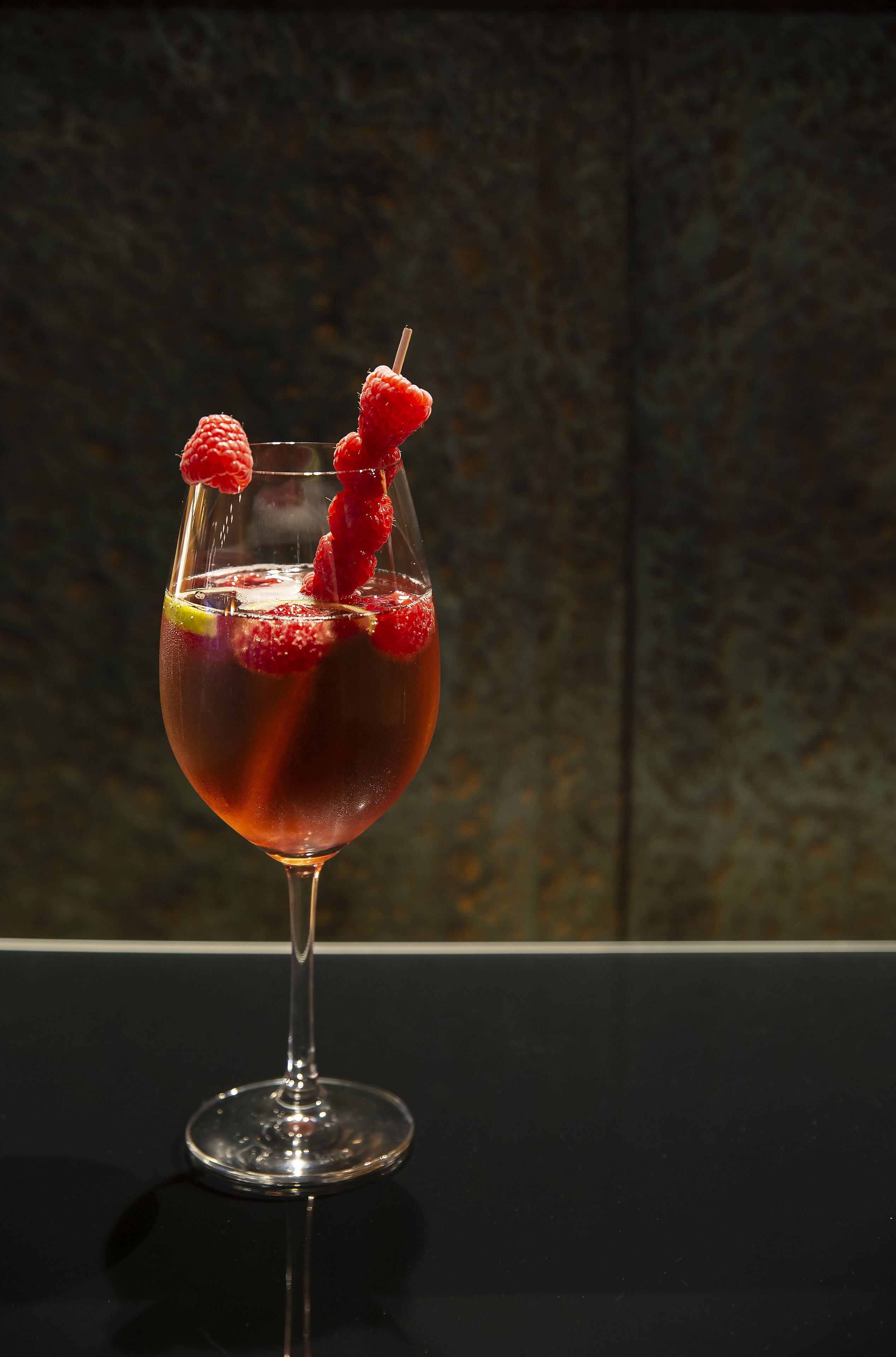 A cocktail glass with a dark liquid, garnished with raspberries and a lemon slice, on a reflective surface.