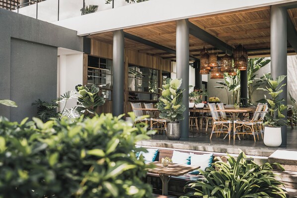 Un área de comedor al aire libre con mesas y sillas de madera, rodeada de grandes macetas con plantas.