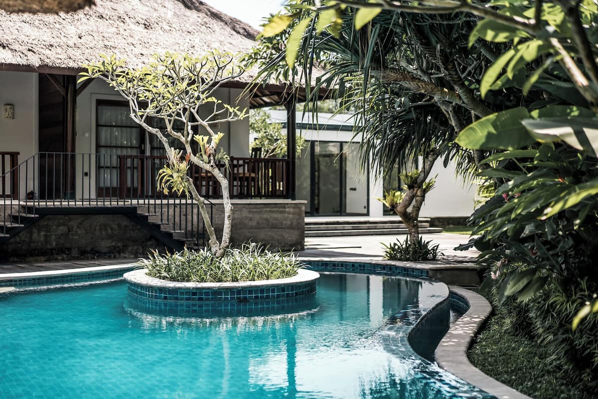 A swimming pool with a tiled edge, surrounded by lush greenery and a thatched-roof building.
