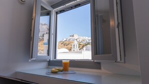 A room with a window overlooking a hill with white buildings, a glass of orange juice, and a banana on the windowsill. - Bedspot Apartments Astypalaia (Astypalaia)