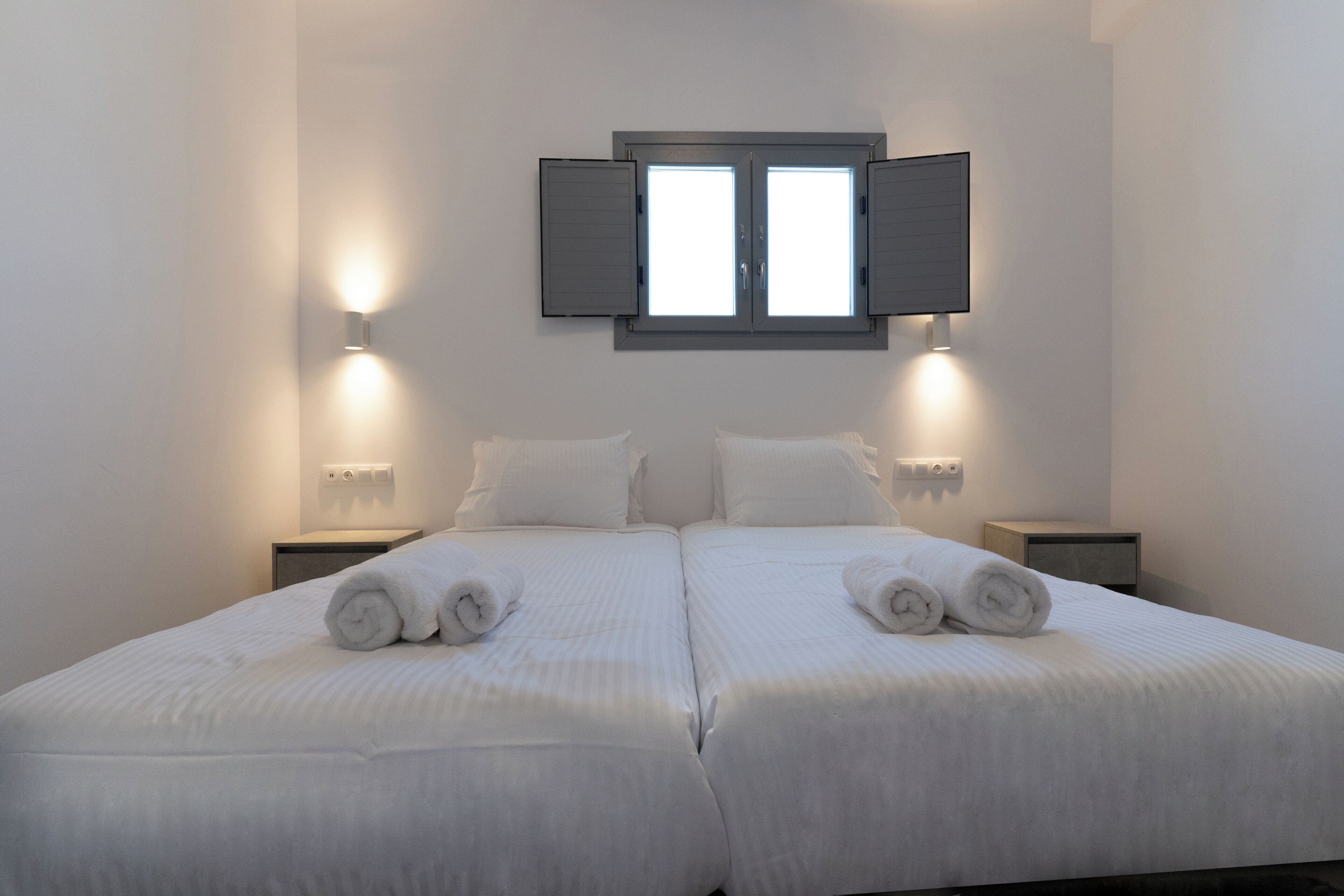 Una cama doble con dos almohadas, una ventana con persianas, lámparas de noche y una mesita de noche.