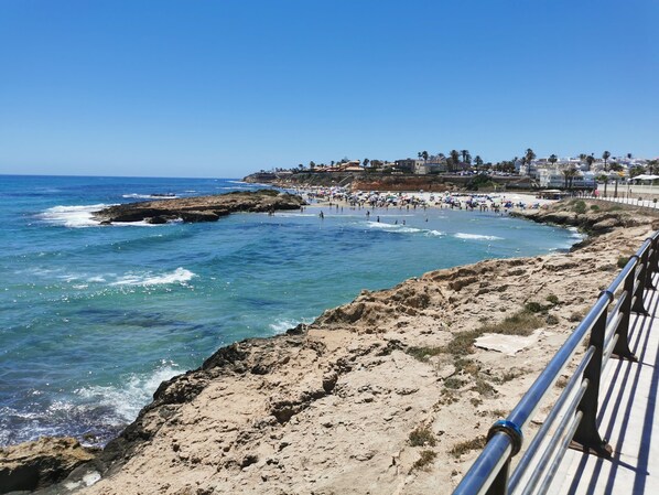 A coastal beach scene with rocky terrain, a clear blue sky, and a promenade with a railing.