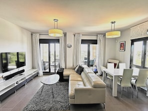A modern living room with a sofa, TV, dining table, chairs, and a balcony with curtains.