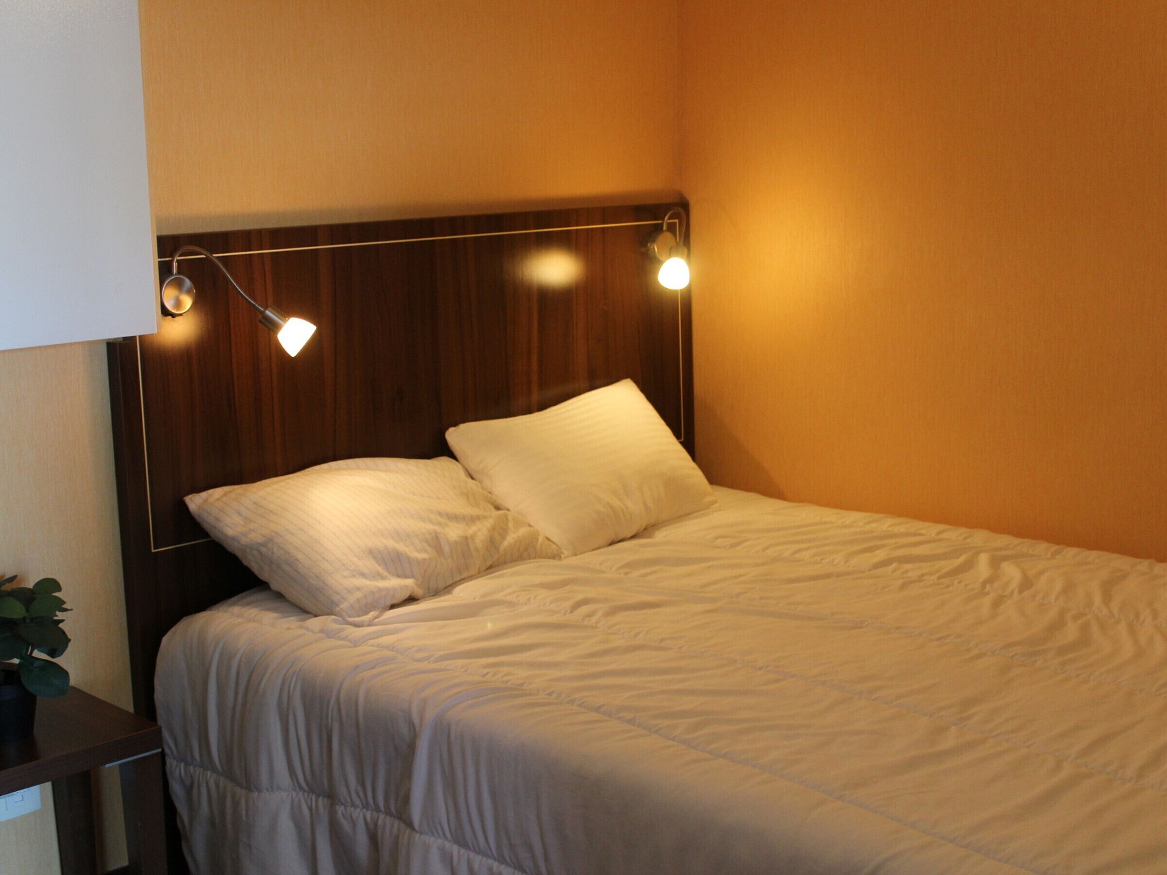 Una cama bien hecha con cabecera, dos almohadas y una mesita de noche con lámpara.