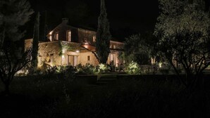 Une grande maison illuminée la nuit, avec une façade en pierre, entourée d’arbres et d’un jardin bien éclairé.