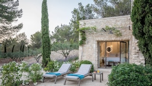 Un bâtiment en pierre avec une porte en verre donnant sur une chambre, deux fauteuils avec des coussins bleus et une petite table sur laquelle est posée une bougie.