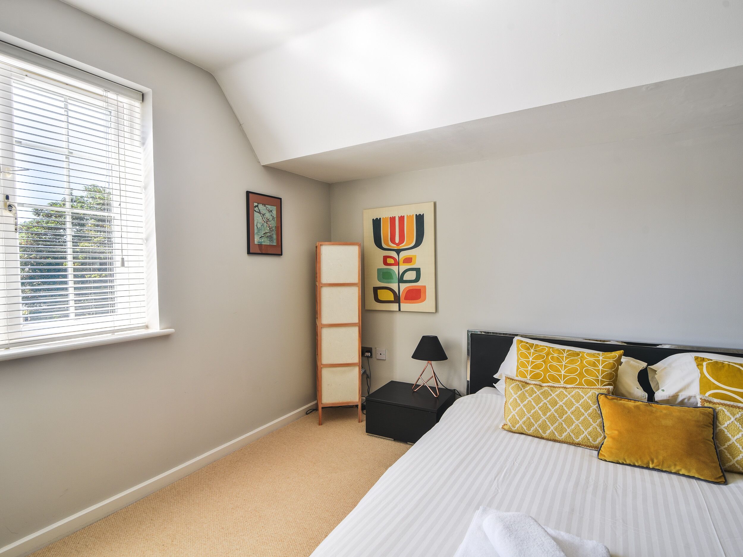 A bedroom with a bed, a window with blinds, a bedside table, a lamp, and a wall with two pieces of artwork.