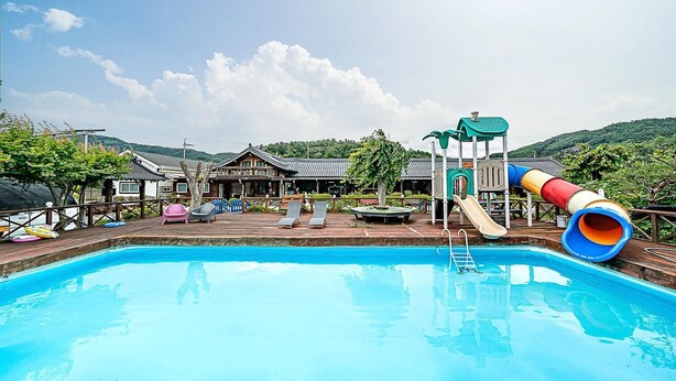 A swimming pool with a slide and a play structure, surrounded by lounge chairs and a traditional building. - Gapyeong Acimpunggeongpension (Gapyeong)