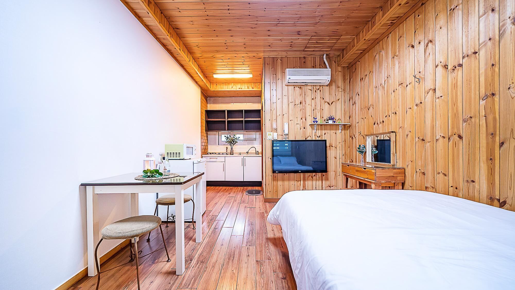 Una habitación compacta con cama, una mesita y una silla, kitchenette y televisor montado en la pared.