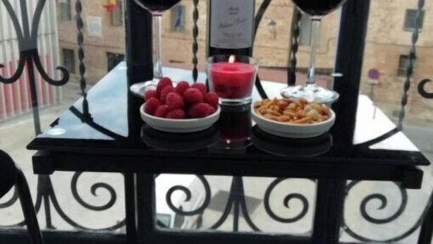 A table with two wine glasses, a bottle of Protas wine, a bowl of raspberries, a bowl of nuts, and a candle on a balcony with a wrought iron railing.