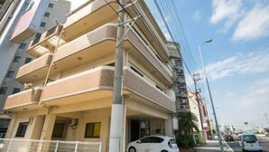 A multi-story building with a parking area and a white car parked in front. - Wing Mihama Beach (Chatan)