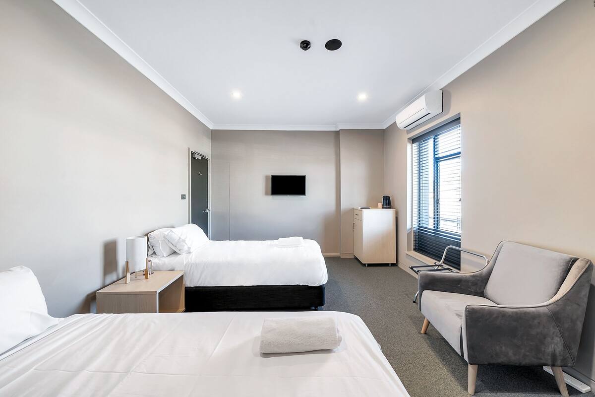 Habitación de hotel con dos camas, un televisor de pantalla plana, un escritorio, una silla y una ventana con persianas.