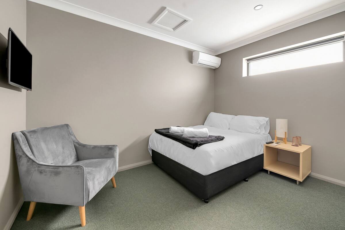 Un dormitorio moderno con una cama, un sillón gris, una mesita de noche con una lámpara y un equipo de aire acondicionado.