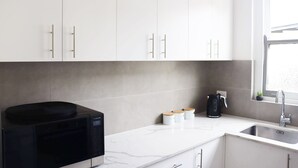 A modern kitchen with white cabinets, a black microwave, a coffee maker, and a window with a view. - Illinois Hotel (Five Dock)