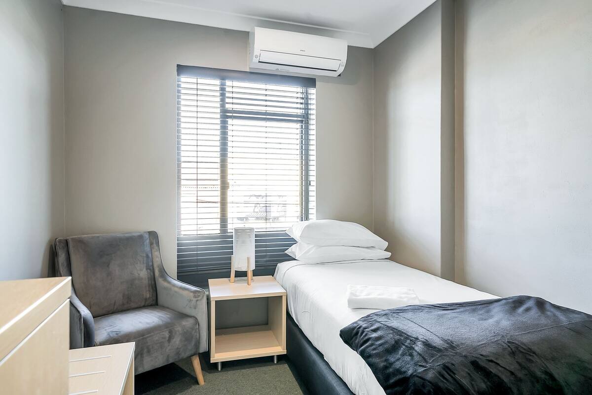 Una habitación de hotel con cama, una silla, una mesita de noche y un ventanal con persianas.