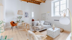 A spacious living room with a wooden ceiling, a grey sofa, a white dog, and a dining area with a table and chairs. - Casa Lazzaro Contemporary Ground Floor Apartment (Lucca)
