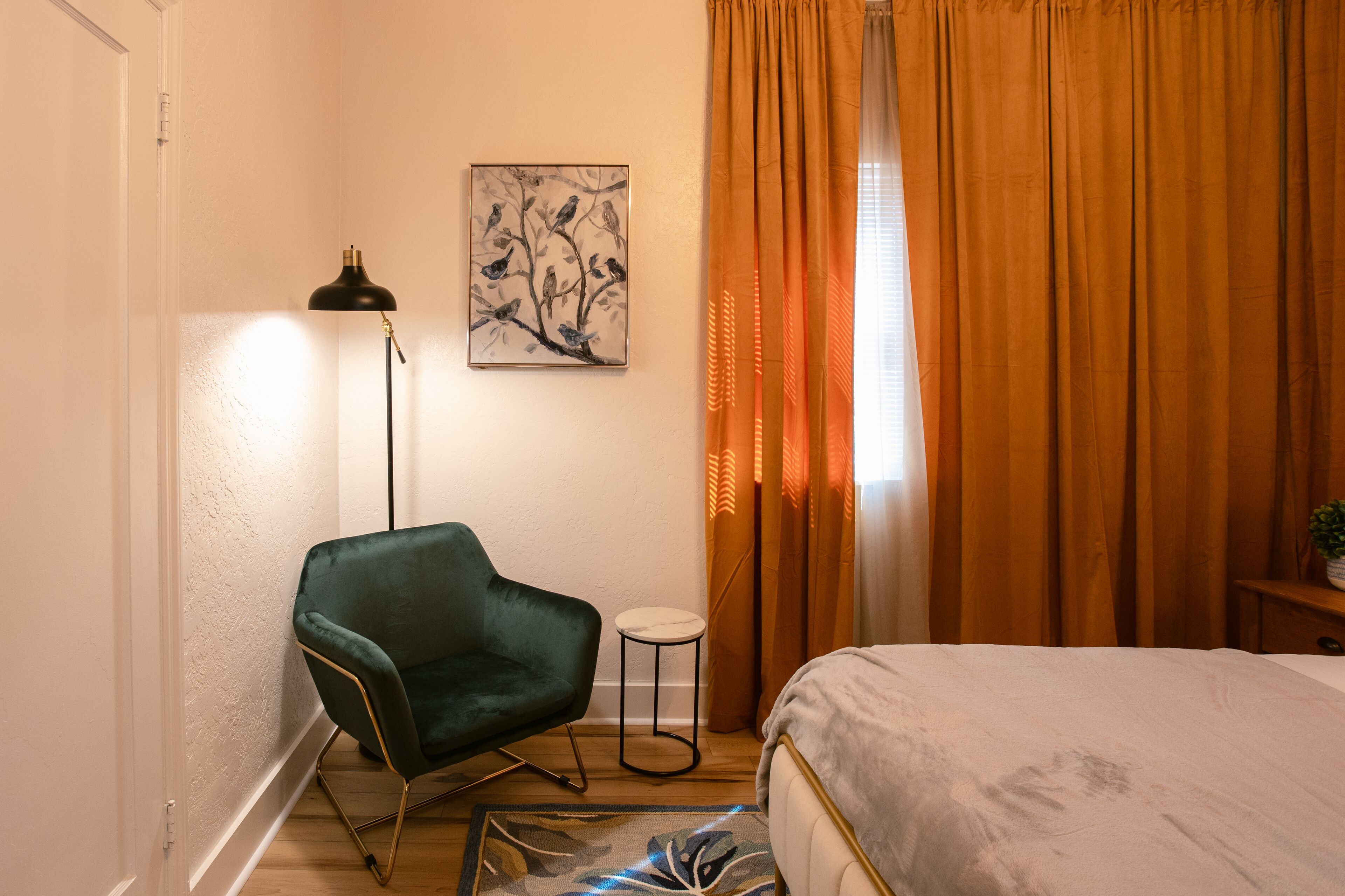 A bedroom with a bed, a green chair, a floor lamp, a painting, and orange curtains.