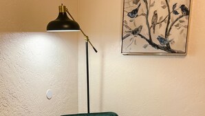 Ett rum med en mörkgrön sammetsfåtölj, en golvlampa, ett litet runt bord och en tavla med fåglar på väggen.