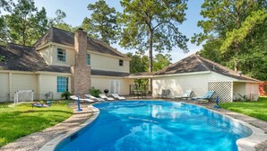 A residential swimming pool with a house in the background, surrounded by trees and a grassy yard.