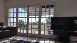Un salon comprenant un canapé, un téléviseur et une porte-fenêtre coulissante donnant sur un balcon avec vue sur des palmiers.