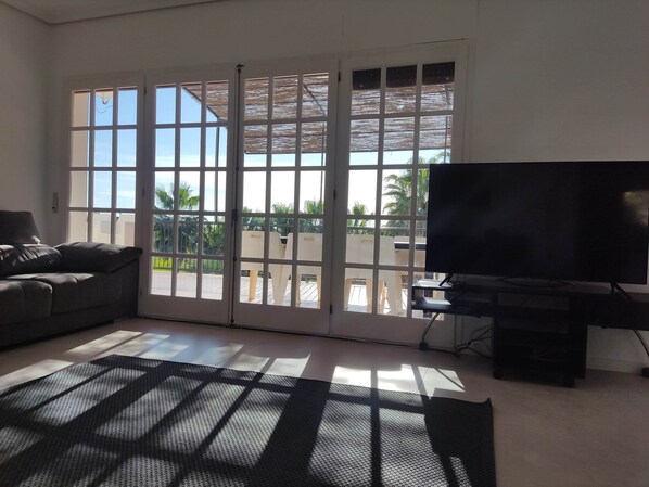 A living room with a sofa, a TV, and a sliding glass door leading to a balcony with a view of palm trees.