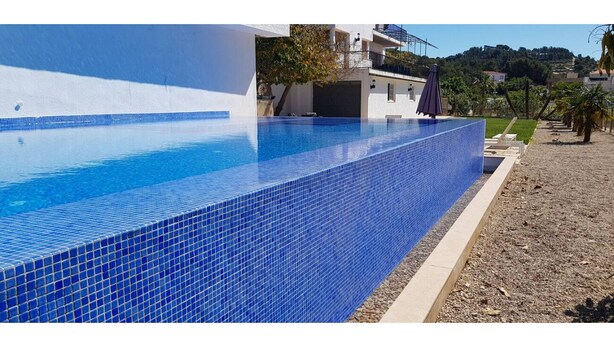 Une piscine rectangulaire aux carreaux bleus, entourée d’une plage de galets et d’un bâtiment blanc doté de balcons.