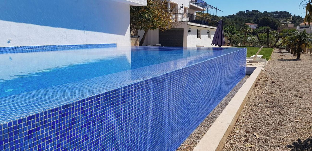 A rectangular swimming pool with blue tiles, surrounded by a pebble beach and a white building with balconies.