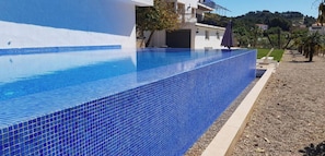 A rectangular swimming pool with blue tiles, surrounded by a pebble beach and a white building with balconies.